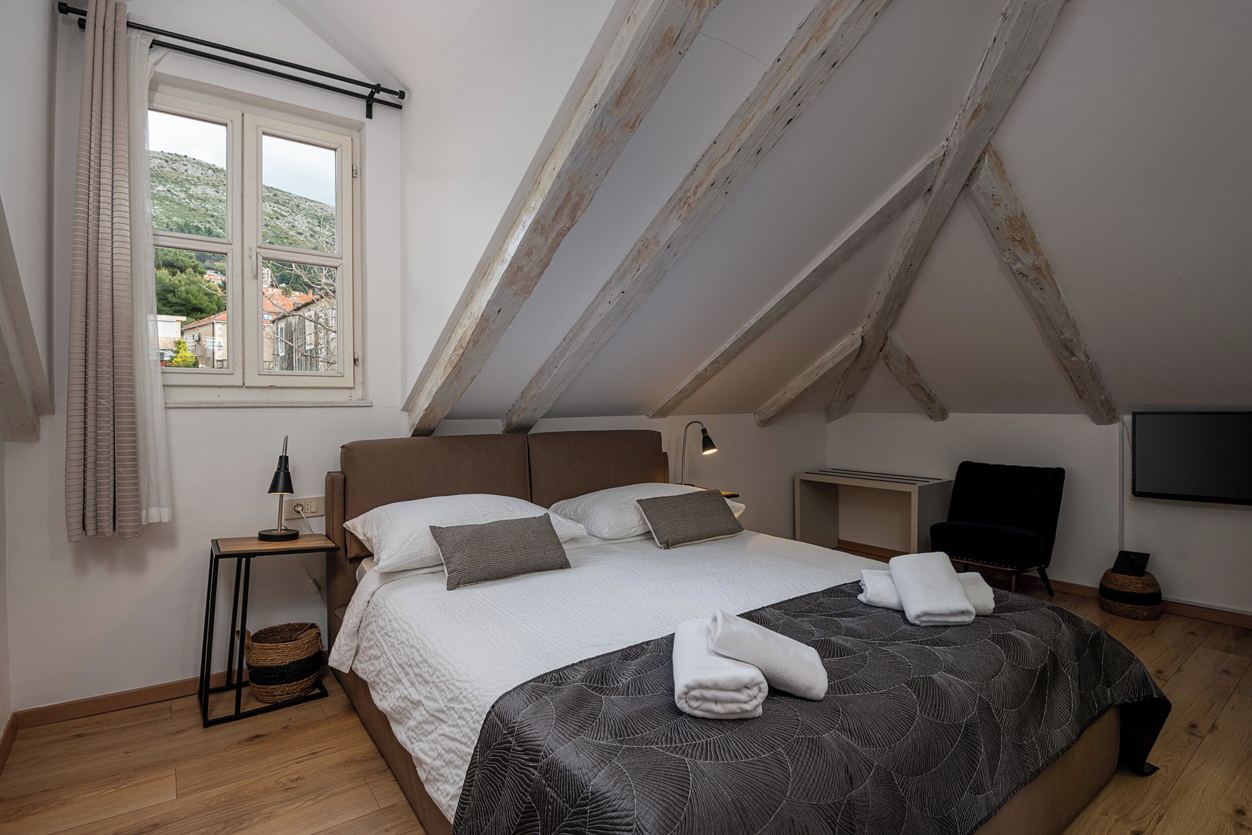 Una camera da letto con soffitto spiovente, un letto con copripiumino decorato, un comodino con lampada e vista sull'esterno attraverso una finestra.