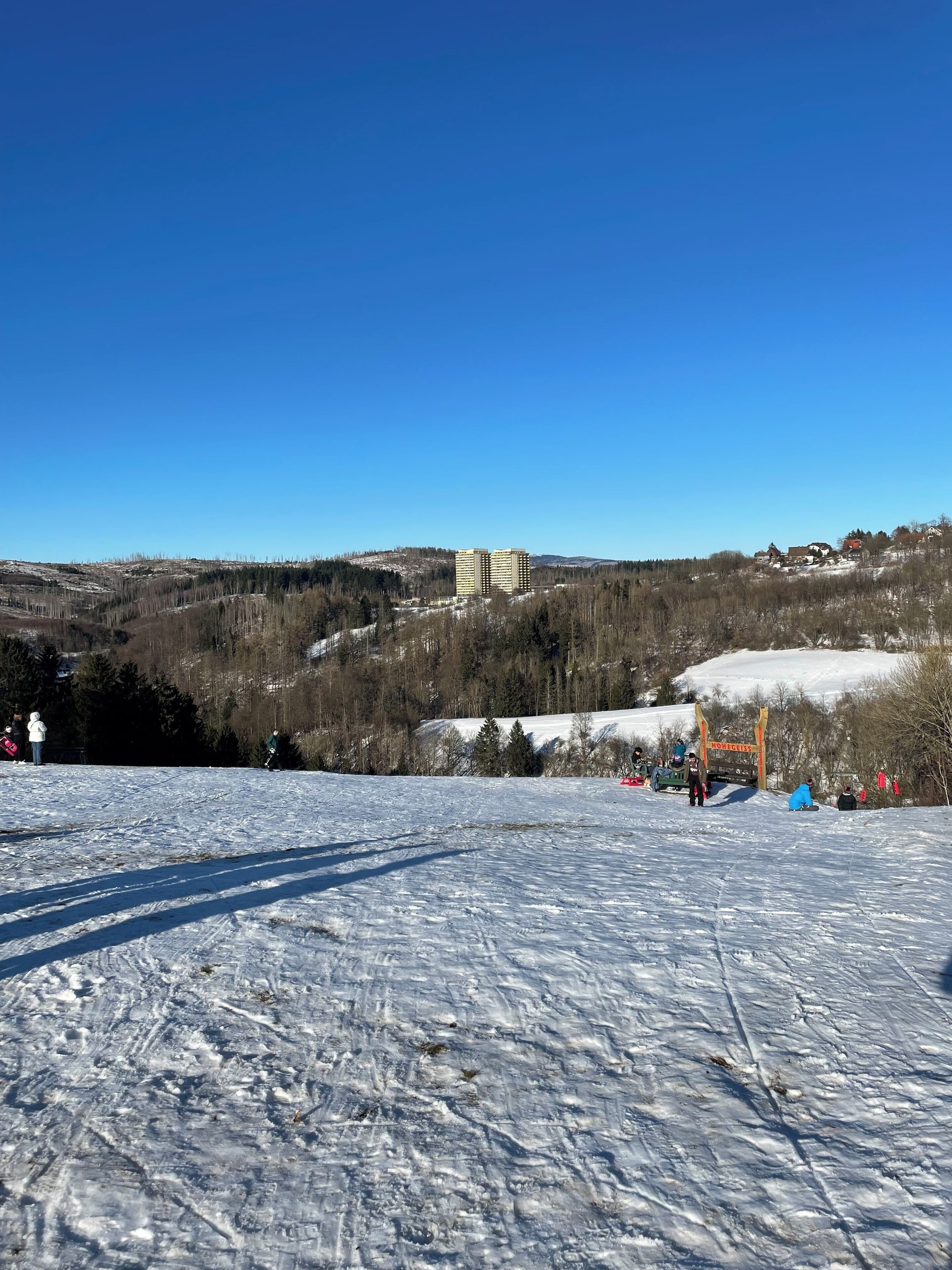 A snowy ski slope with a clear blue sky, a lodge in the background, and a sign reading "Honeigess".
