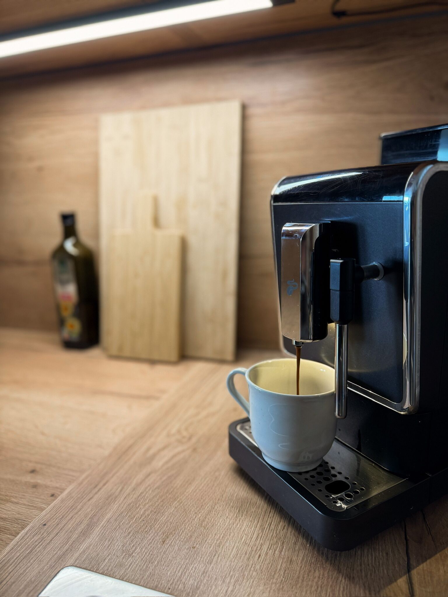 A coffee machine on a countertop with a cup underneath, a bottle in the background, and a wooden panel.