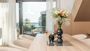 A modern room with a wooden table, a vase of flowers, a wine glass, and a bottle. A large window offers a view of the sea and palm trees.