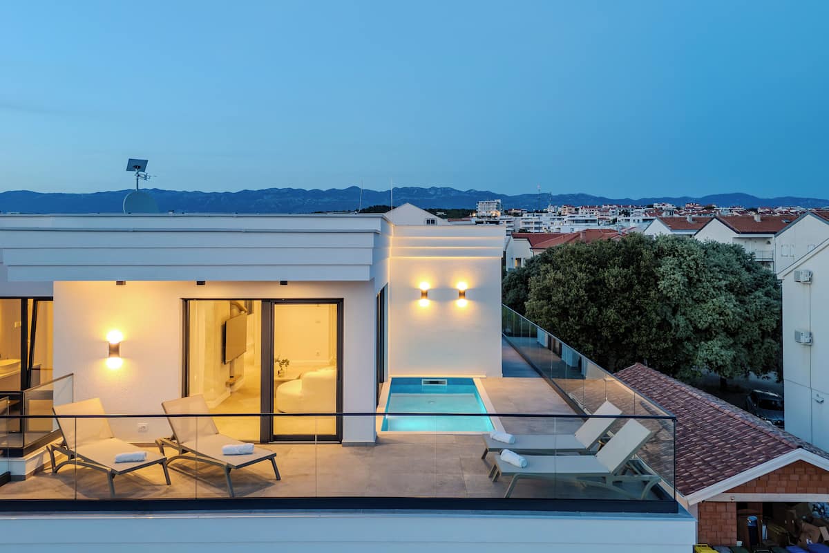 Una azotea moderna con piscina, sillas de descanso y barandilla de cristal con vistas a un paisaje urbano y montañas al fondo.