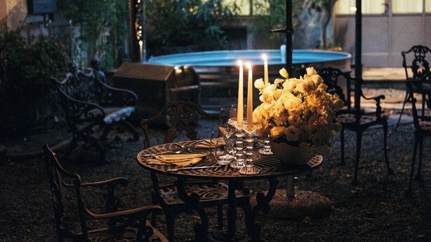 Un espacio de comedor al aire libre con mesa, sillas, una sombrilla, velas y un arreglo floral.