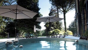 Una piscina con dos sombrillas grandes, rodeada de árboles y mobiliario de exterior.