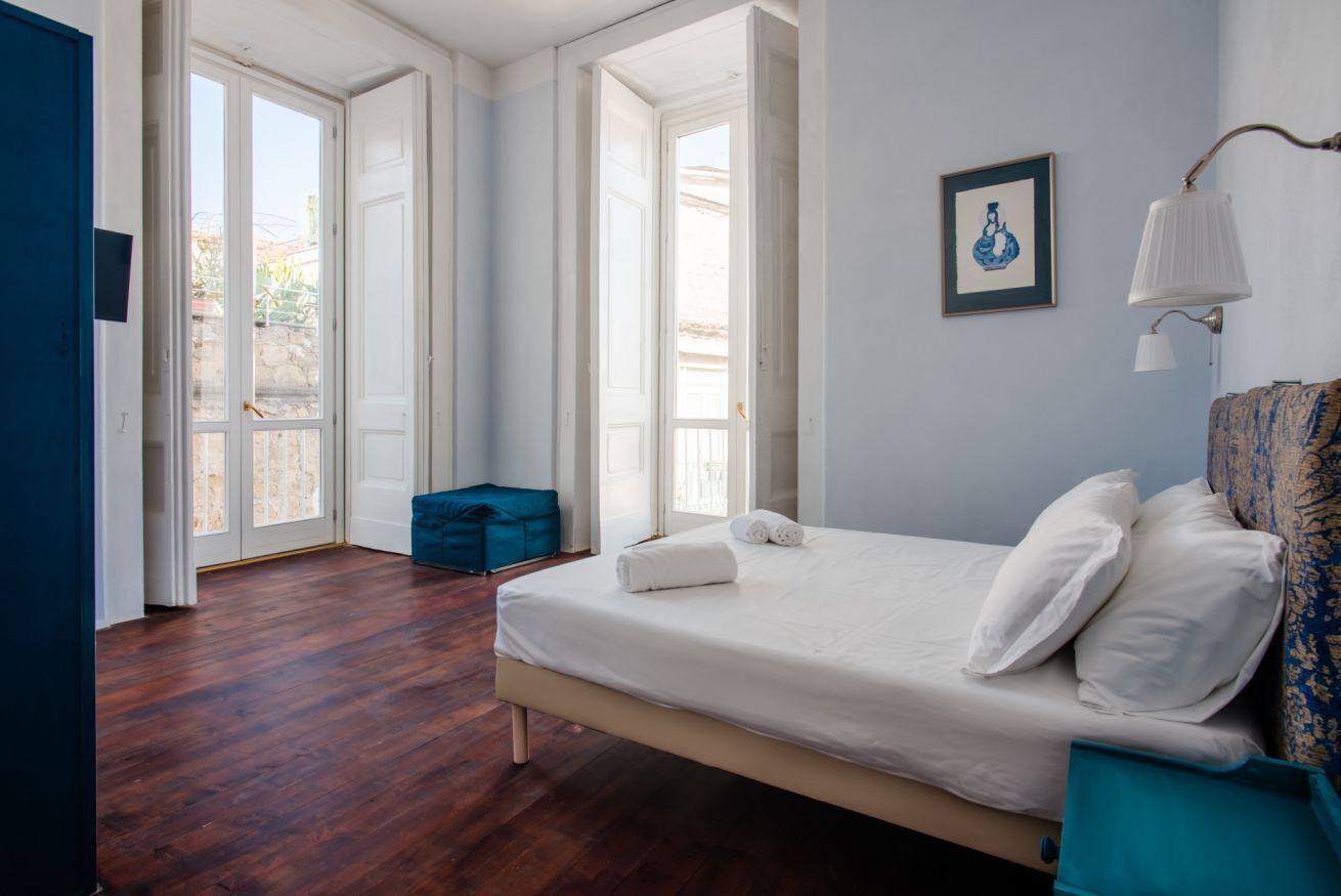 Une chambre avec un grand lit, un pouf bleu et une vue sur l’extérieur, visible à travers une porte vitrée.