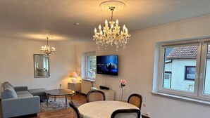 A modern living room with a dining area, wooden flooring, a grey sofa, a chandelier, a TV, and a window with a view of a house.