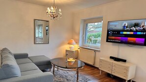 A modern living room with a grey sofa, a glass coffee table, a chandelier, a TV mounted on the wall, and a window with a view of greenery.