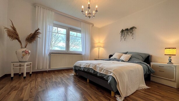 A bedroom with a bed, bedside tables, a radiator, a window with curtains, a chandelier, and a lamp.
