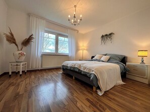 A bedroom with a bed, bedside tables, a radiator, a window with curtains, a chandelier, and a lamp. - Watt'n Traum EG - Your retreat by the North Sea (Wurster Nordseeküste)