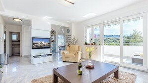A modern living room with a flat-screen TV, a beige sofa, a coffee table with a bottle and glass, and large windows with a view of greenery. - Apartment Aqua-Juric (Crikvenica)