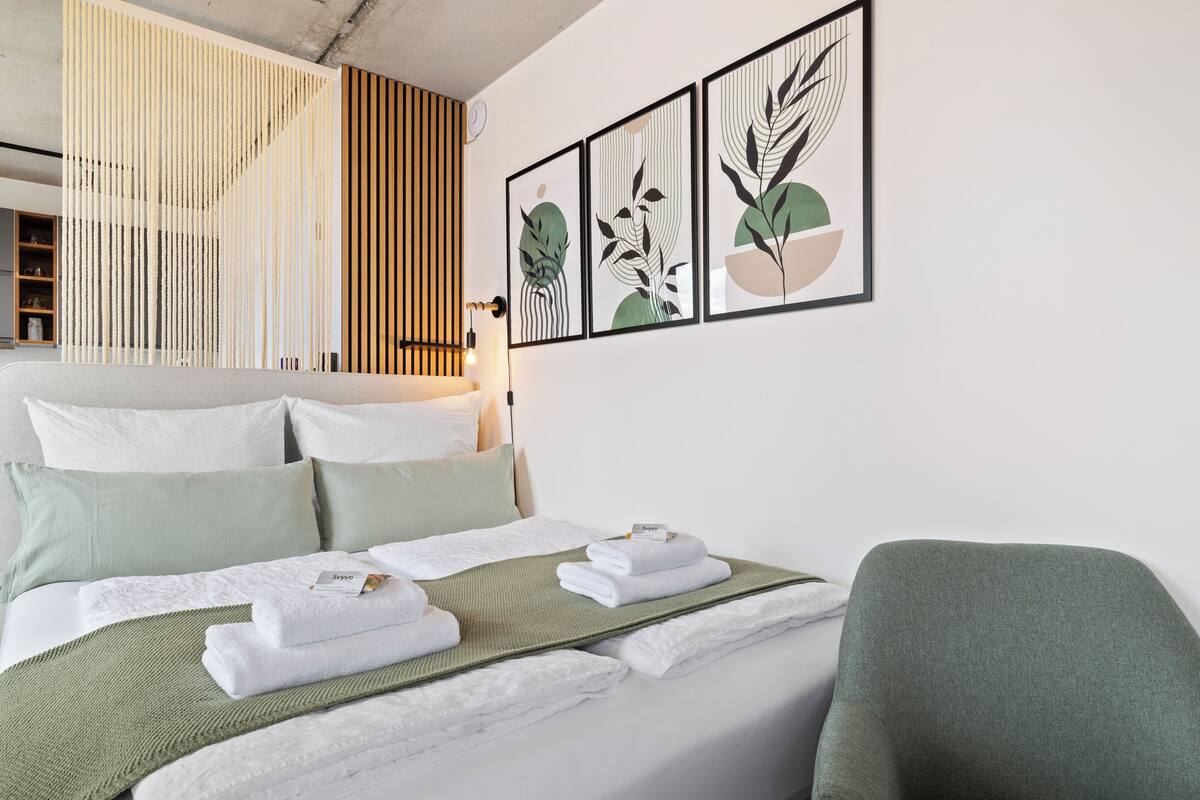 Uma cama bem arrumada com lençóis brancos e um cobertor verde, uma poltrona verde e uma parede com impressões botânicas emolduradas.