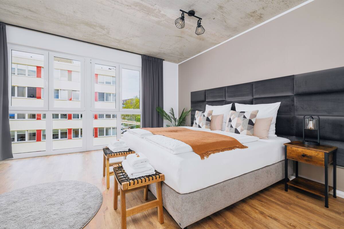 Quarto moderno com uma cama grande, mesas de cabeceira de madeira, um tapete cinzento e vista para edifícios através de amplas janelas.