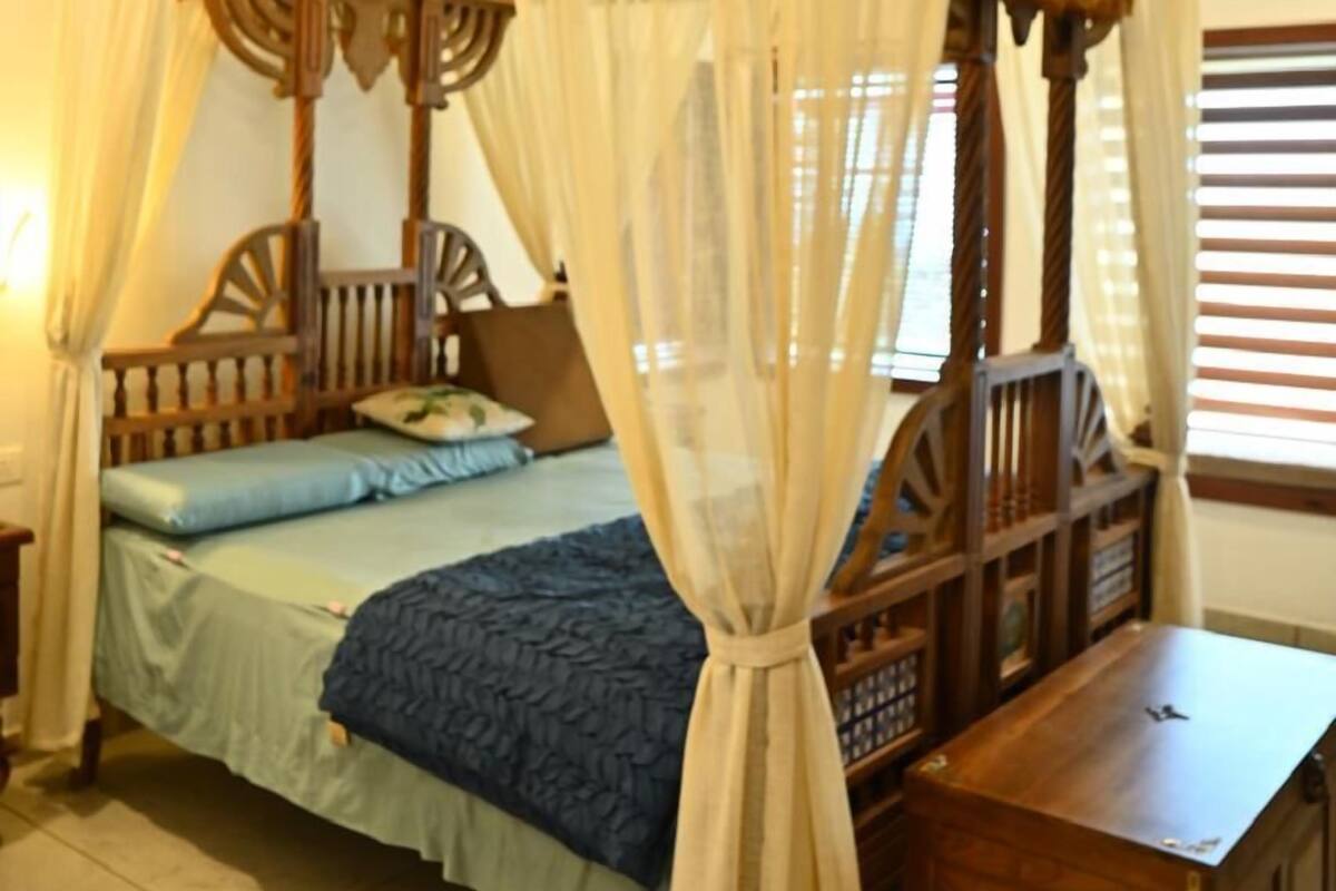 A four-poster canopy bed with a wooden frame and white drapes, a wooden nightstand, and a bench at the foot of the bed.