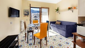 A living room with a blue sofa, a dining table with chairs, a TV mounted on the wall, and a view of buildings through a glass door. - Lovely and typical apartment in the center of Sorrento (Campania)