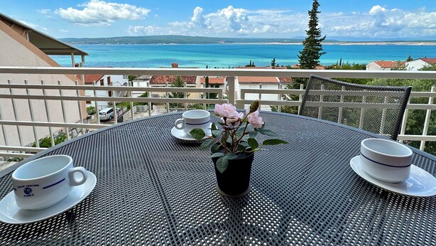 Un balcón con una mesa preparada para dos personas, con vistas a la costa y un cielo despejado.