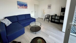 Una sala de estar moderna con un sofá azul, una mesa de centro de vidrio y una silla negra. Hay un cuadro de un paisaje urbano en la pared y una fotografía enmarcada de un edificio.