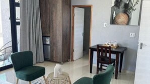 Ein modernes Hotelzimmer mit einem Bett, zwei grünen Sesseln, einem kleinen Tisch, einem Esstisch, einem Holzschrank und einem großen Fenster mit Vorhängen.