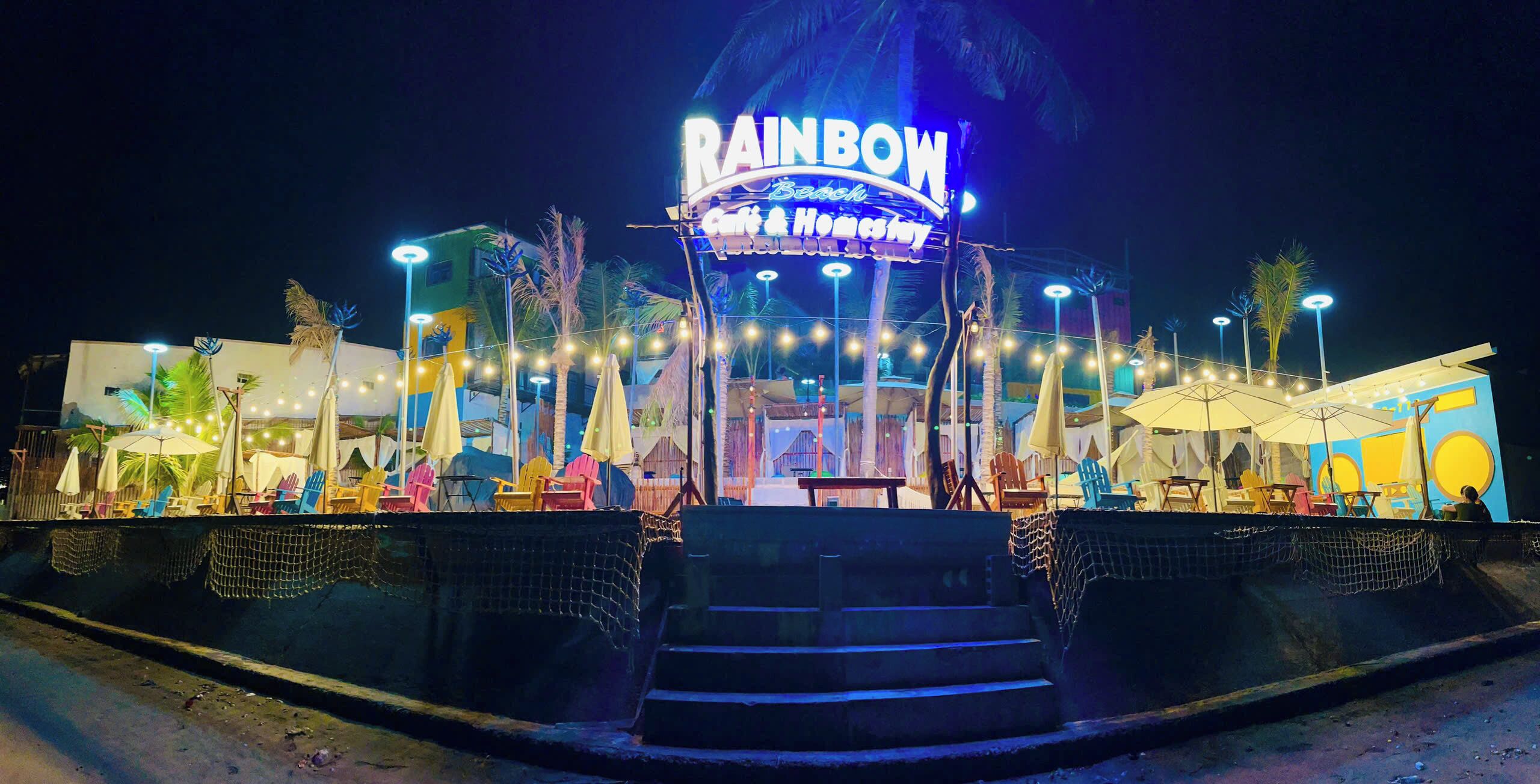 A nighttime view of a beach-themed restaurant named "Rainbow Beach Cafe & Home Stay" with outdoor seating and palm trees.