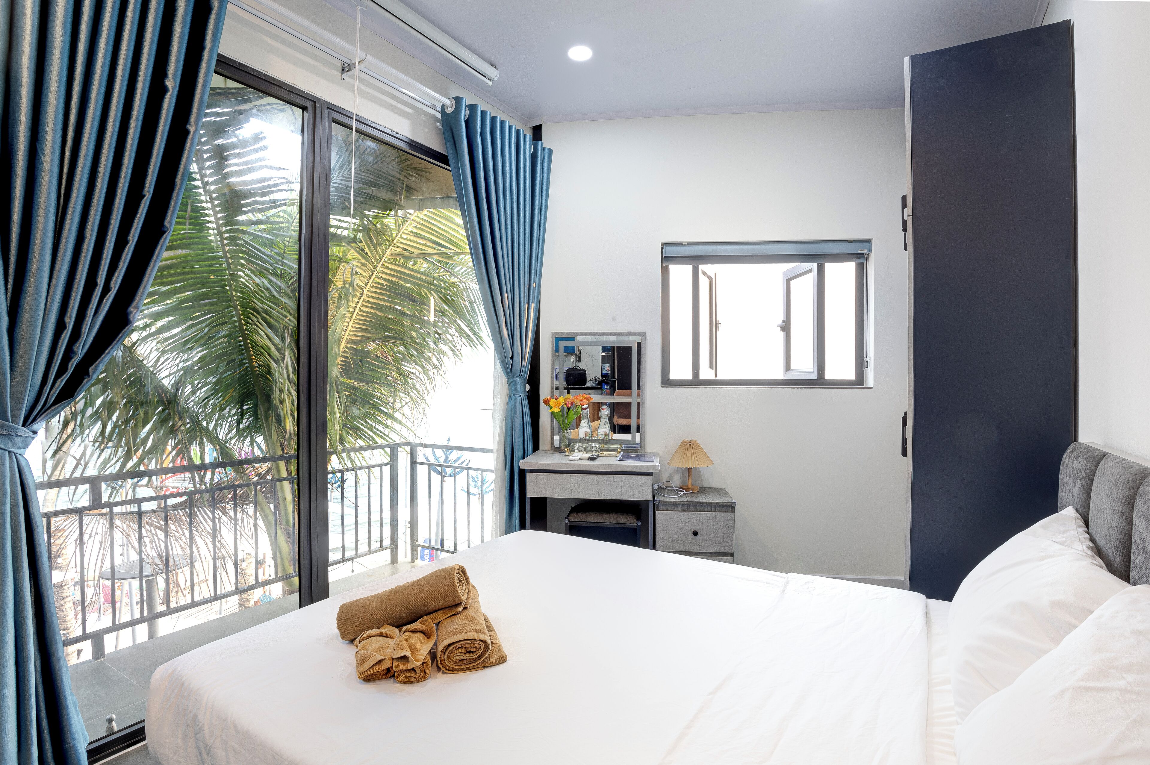 Habitación de hotel con una cama grande, un escritorio con espejo y vistas a palmeras a través de un ventanal amplio.
