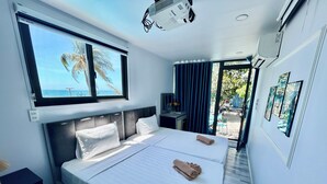Habitación de hotel con cama, escritorio, ventana que ofrece vistas a palmeras y al mar, y puerta que da a un espacio exterior.