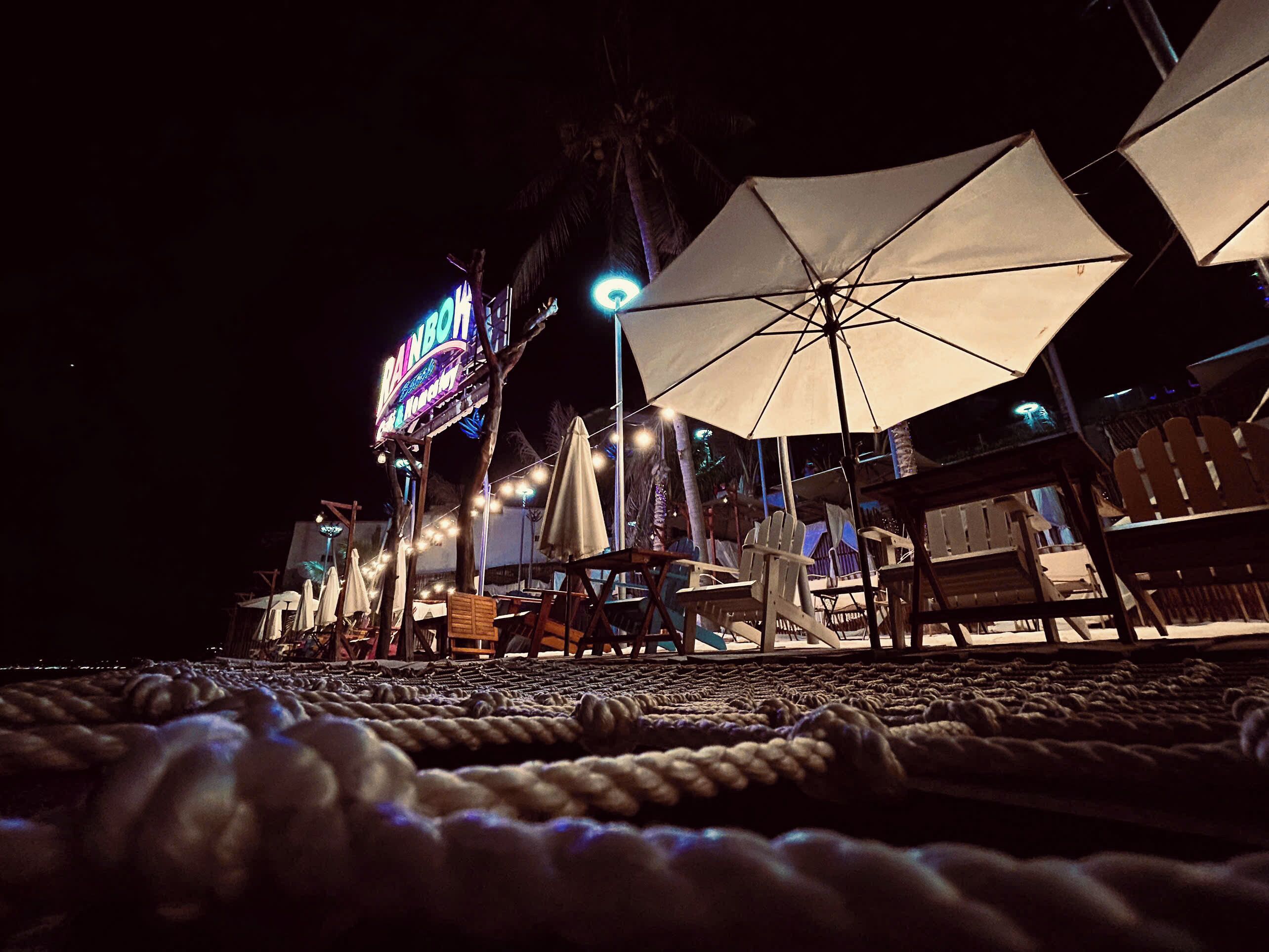 Zona de playa con sombrillas, sillas y un letrero luminoso de neón.