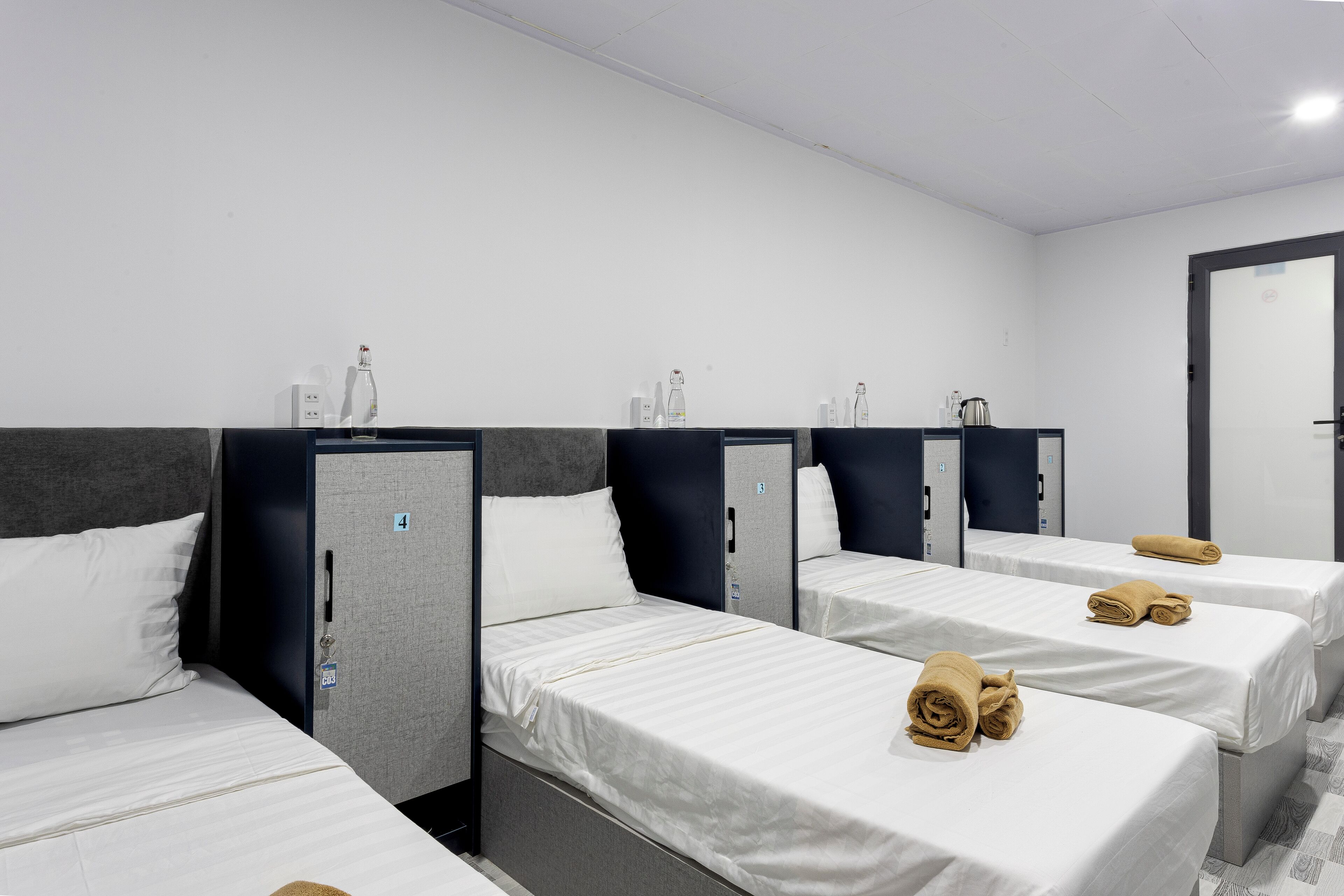 A hotel room with four single beds, each with a pillow and a rolled towel. There are storage units with a keycard lock and a door with a number.