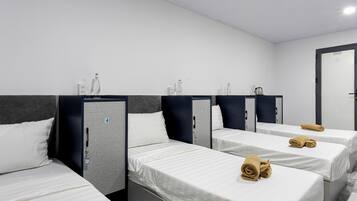 A hotel room with four single beds, each with a pillow and a rolled towel. There are storage units with a keycard lock and a door with a number.