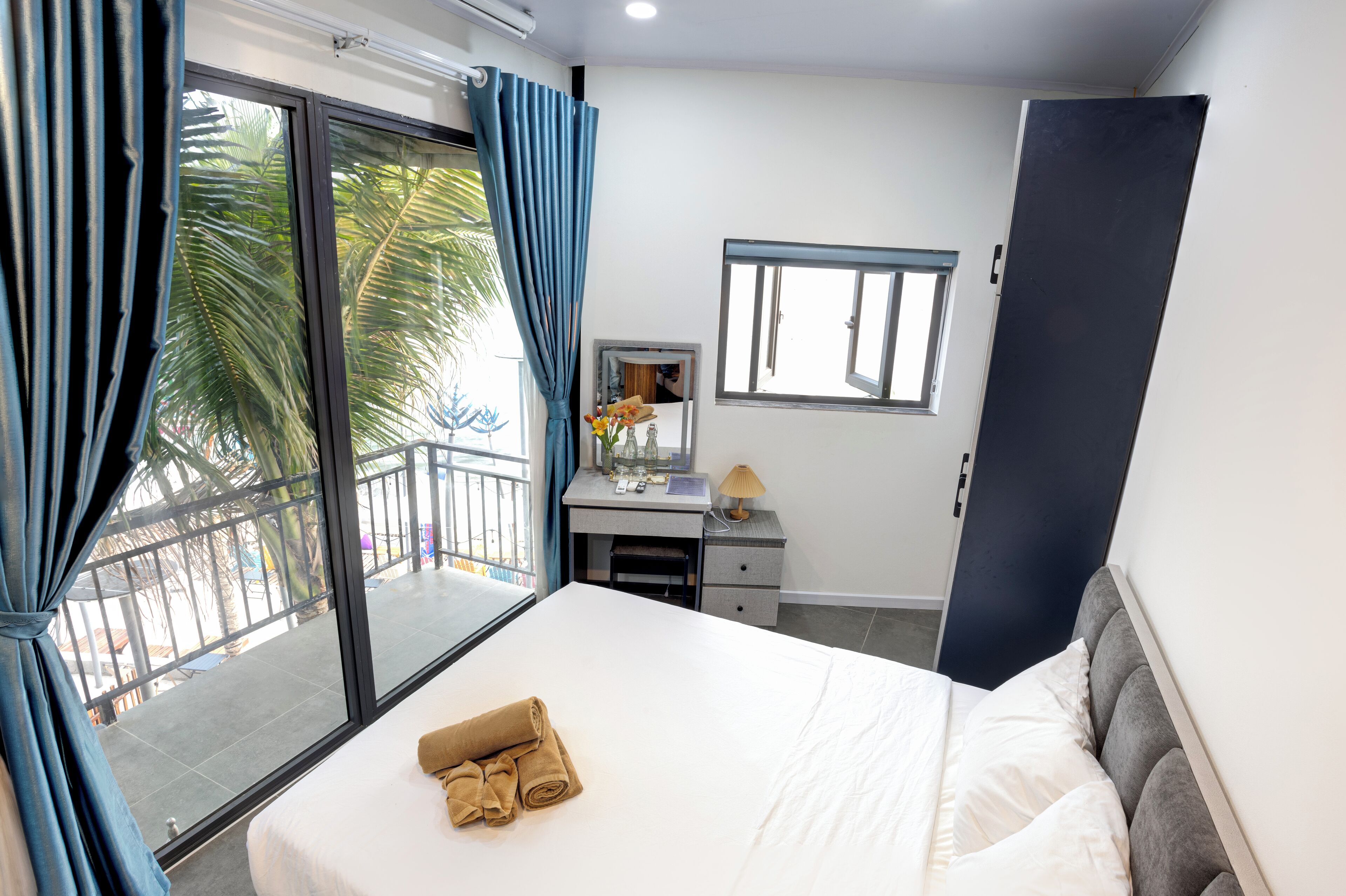 Une chambre d’hôtel avec un lit, un bureau doté d’une lampe, une fenêtre et une vue sur des palmiers.