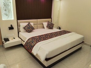 A neatly made bed with a patterned bedspread, two pillows with paisley designs, a bedside table with a phone and a remote control, and a wall-mounted light fixture.