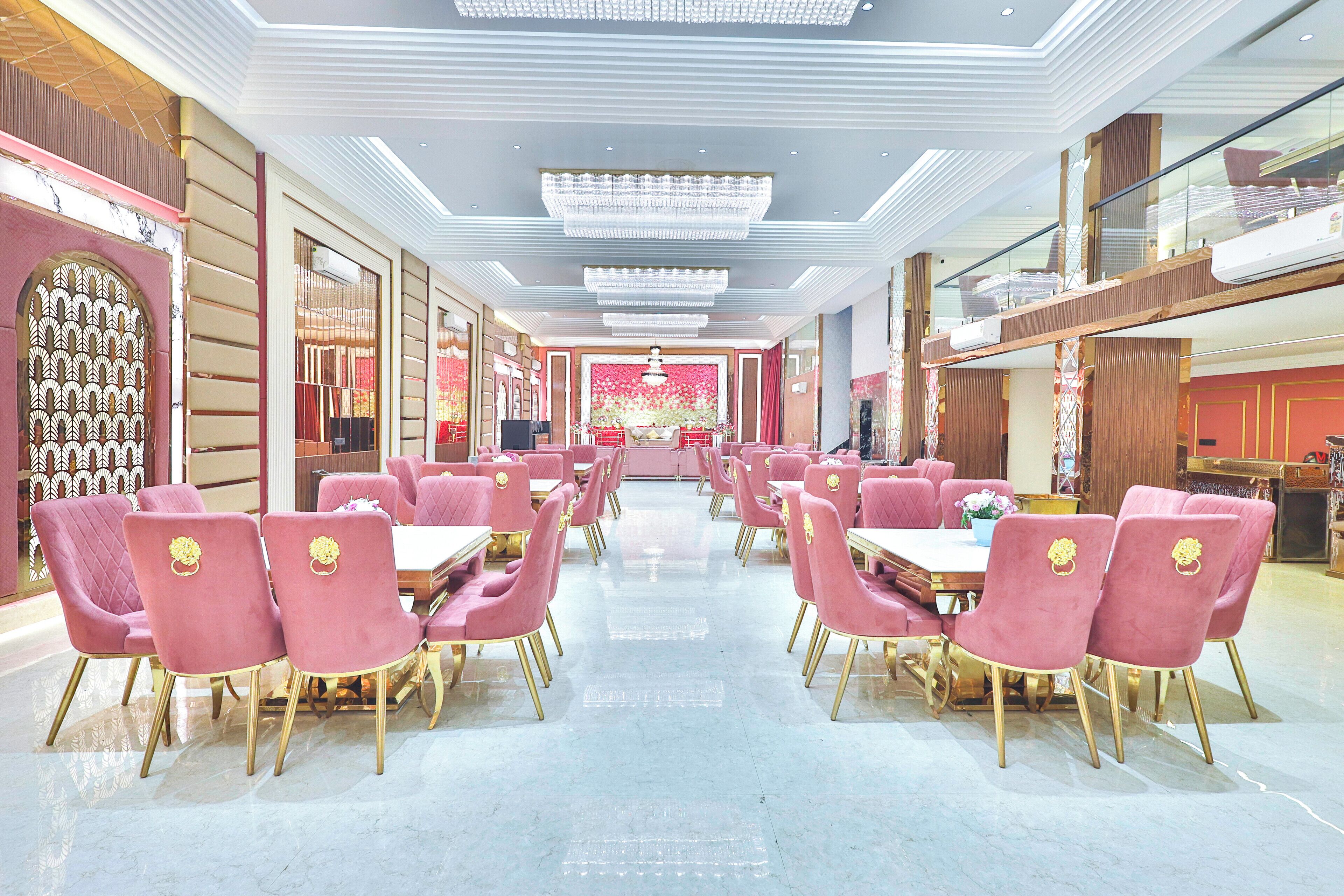 A spacious restaurant with pink chairs and gold accents, featuring a central stage and a modern ceiling design.