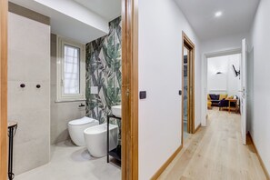 Un baño moderno con inodoro, lavamanos y ducha, que da paso a una sala de estar con sofá y mesa.