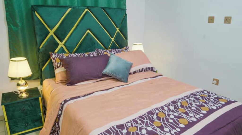 Ein Bett mit einem gemusterten Überwurf, ein grüner Kopfteil mit geometrischem Muster und zwei Nachttischlampen auf einem grünen Nachttisch.