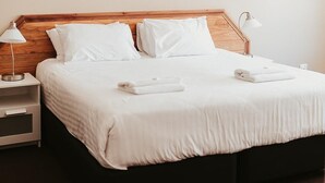 A neatly made bed with white linens and pillows, a wooden headboard, a bedside table with a lamp, and a small nightstand.