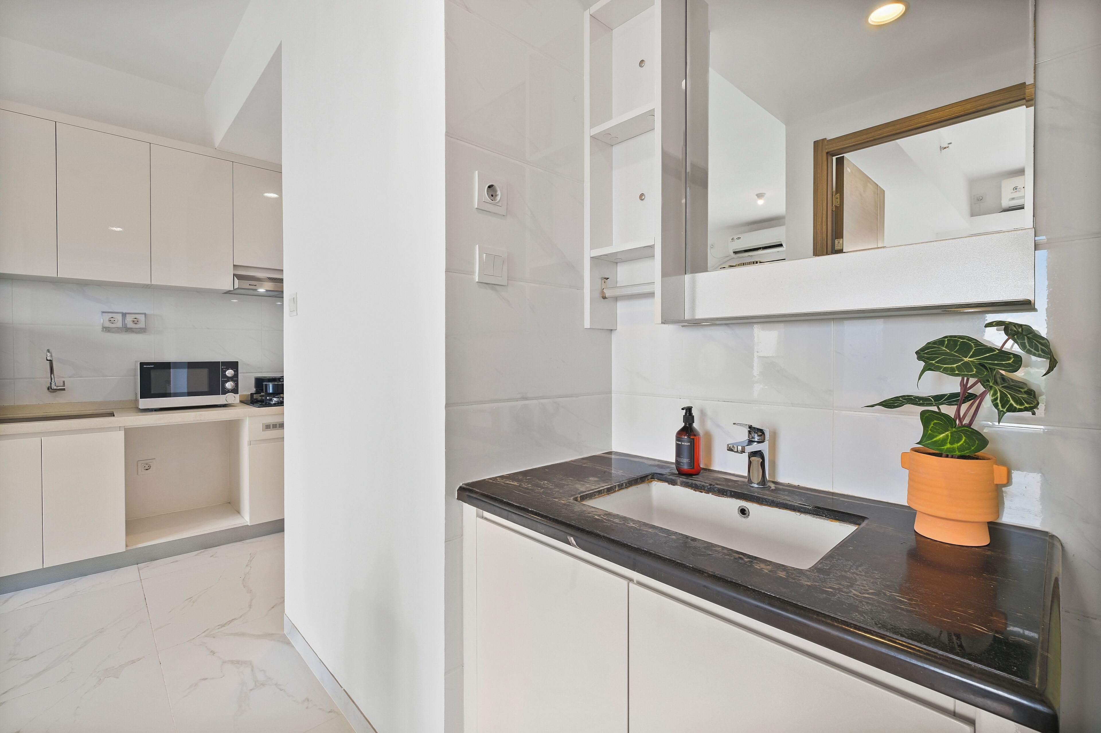 Een moderne keuken met witte kastjes, een zwart aanrecht en een spoelbak. Op het aanrecht staat een magnetron, een plant en een fles.