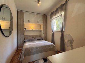 A bedroom with a bed, a bedside table, a mirror, a window with curtains, a ceiling light, and built-in cabinets. - Apartment Lu&Ni (Crikvenica)