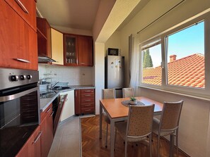 A compact kitchen with wooden cabinets, a built-in oven, a refrigerator, and a dining table with chairs. - Apartment Lu&Ni (Crikvenica)