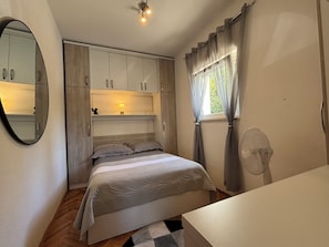 Ein Schlafzimmer mit Bett, Nachttisch, Spiegel, Kleiderschrank, einem Fenster mit Vorhängen und einer Deckenleuchte.