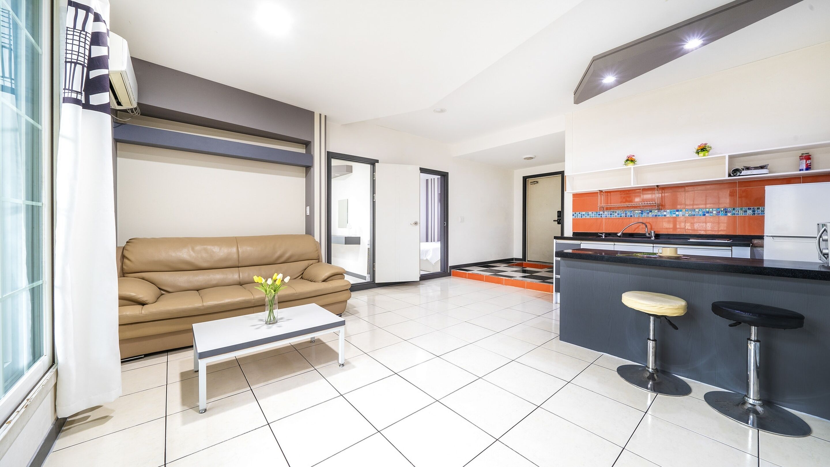 A modern living room with a beige sofa, a coffee table with a vase, and a kitchen area with a black countertop and two stools.