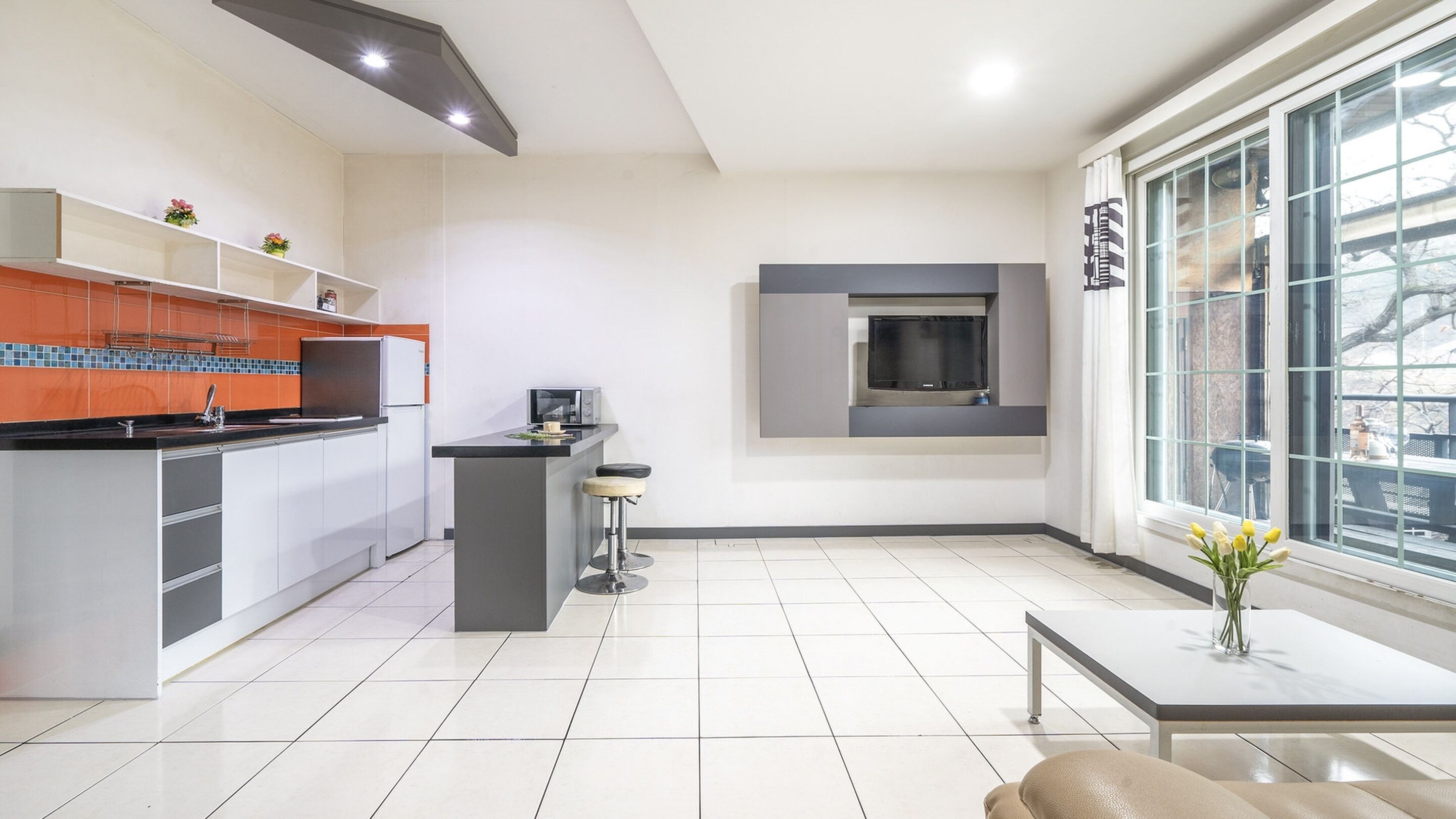 A modern kitchen with white cabinets, a black countertop, and a built-in oven. There is a small island with a sink and a chair, and a large window with a view of the outside.