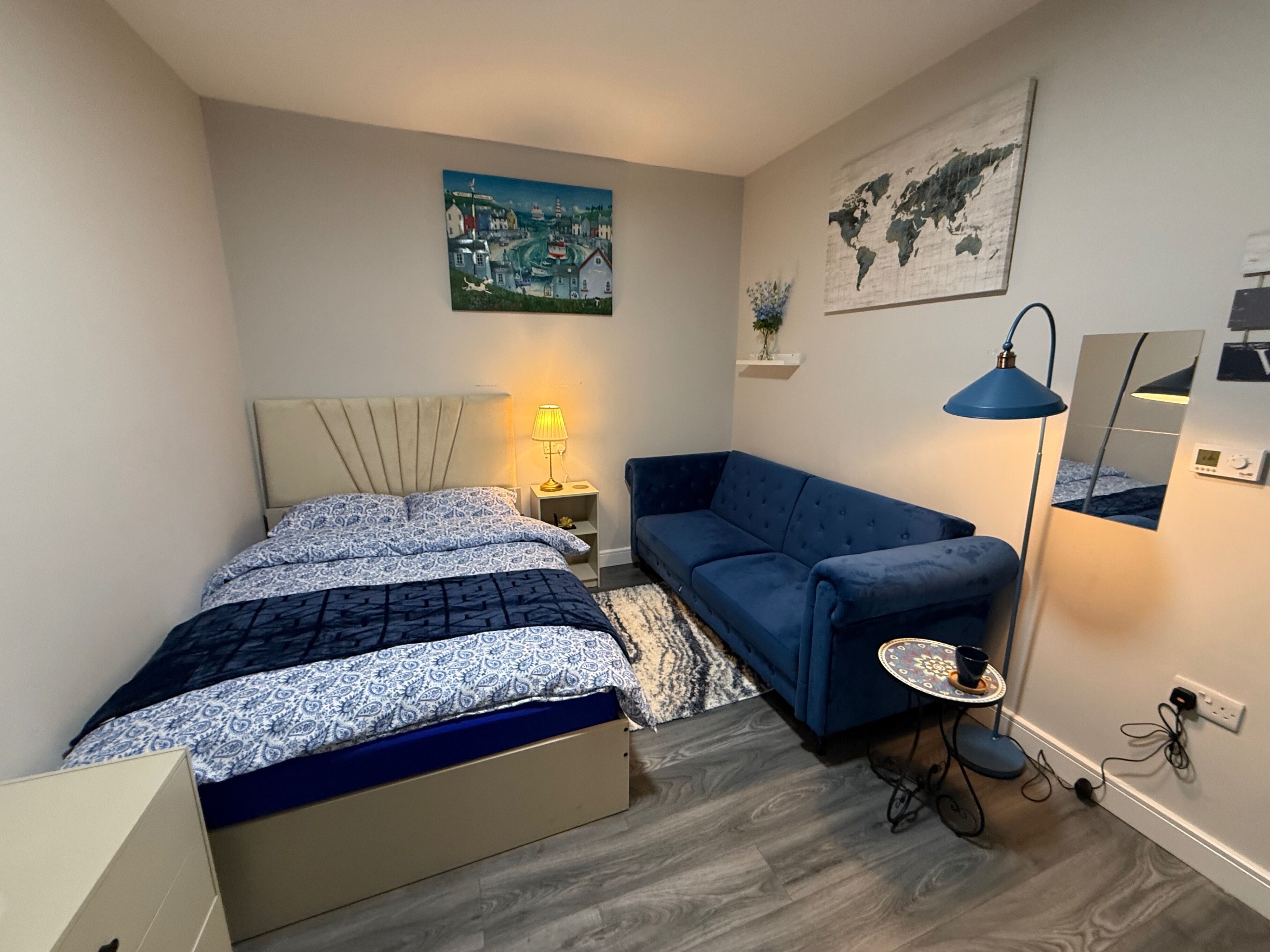 A compact bedroom with a bed, a blue sofa, a small table with a lamp, and a world map on the wall.