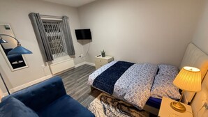 A bedroom with a bed, a blue sofa, a bedside table with a lamp, a TV, a window with curtains, and a radiator.