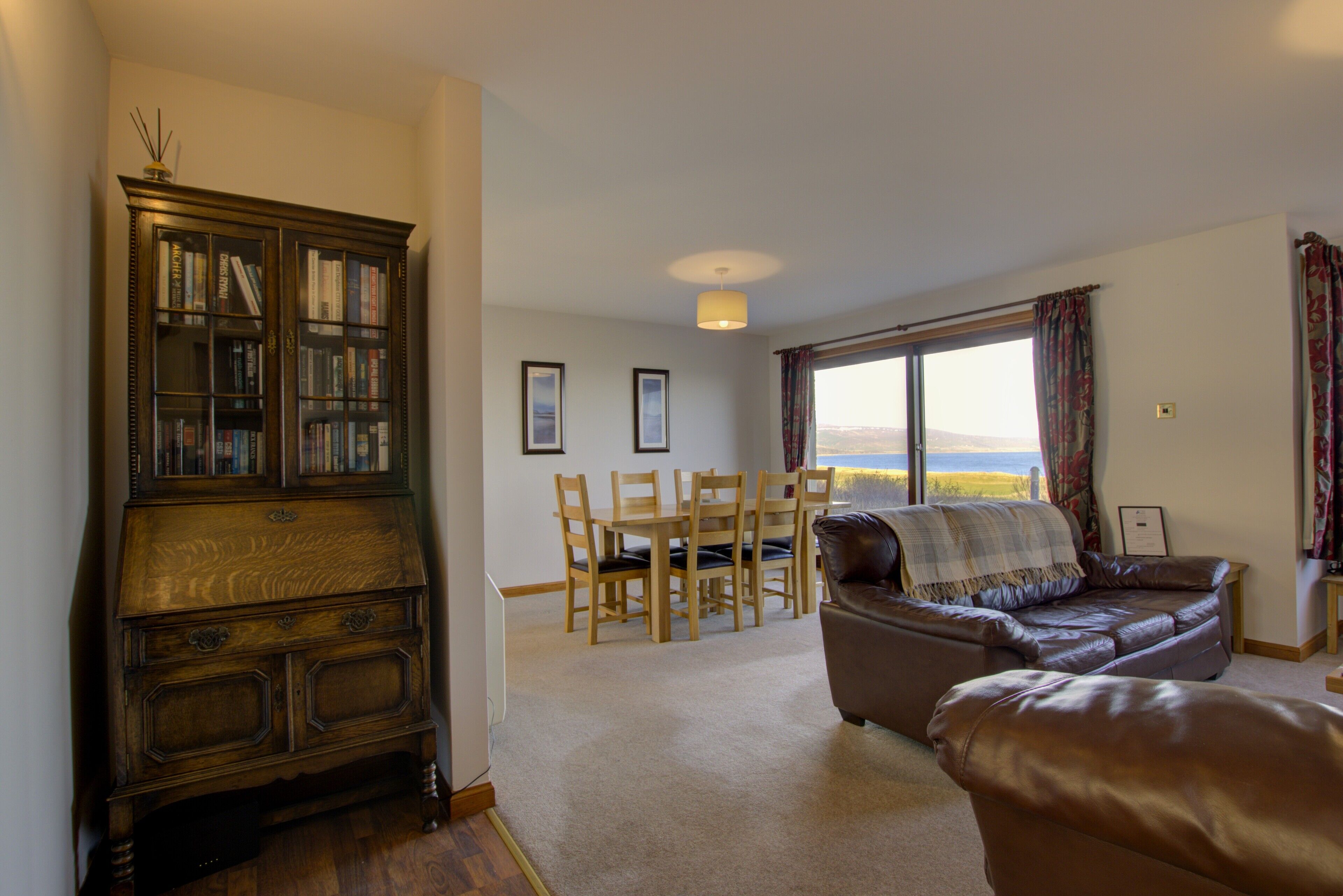 A living room with a wooden cabinet, a brown leather sofa, a dining table with chairs, and a large window with a view.