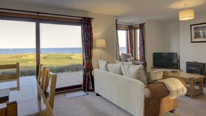 A living room with a sofa, TV, and a table. There is a view of the ocean through large windows. - 2 Links View Brora in Brora (Brora)