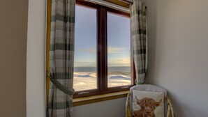 A room with a large window overlooking a beach, a wicker chair with a cushion, and plaid curtains. - 2 Links View Brora in Brora (Brora)