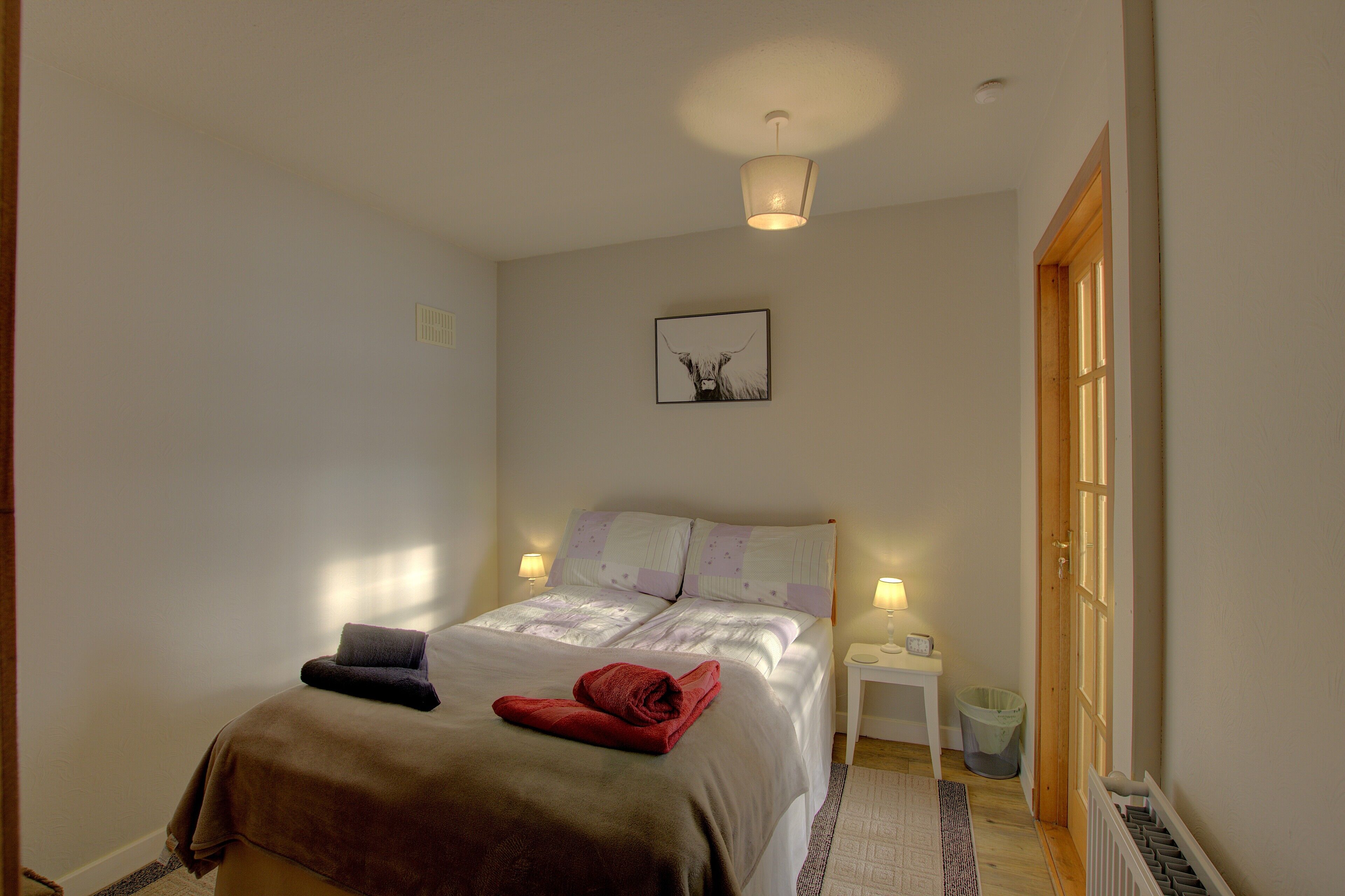 A neatly made bed with a brown blanket and a red towel, a bedside table with a lamp, and a picture on the wall.