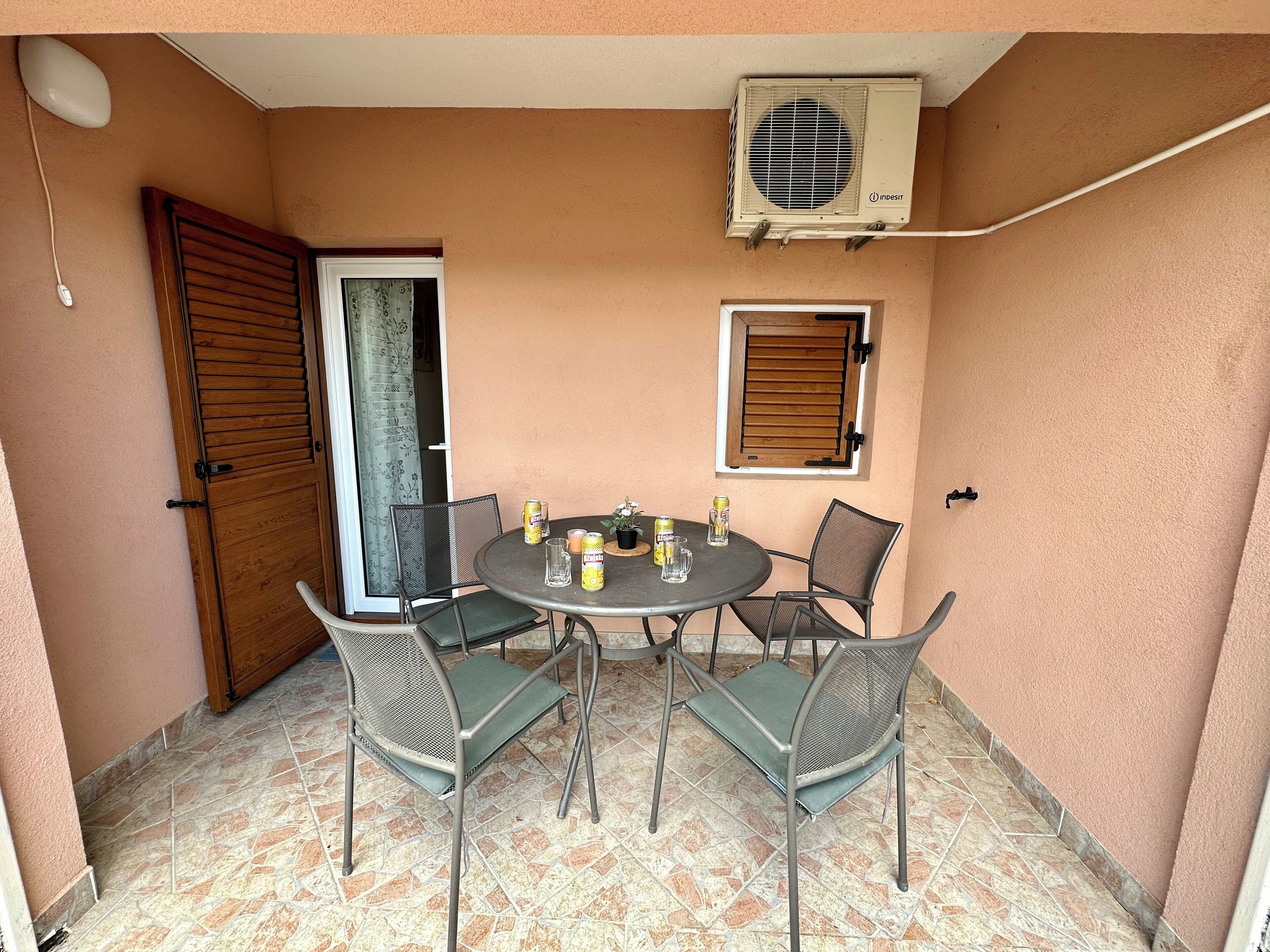 Eine Terrasse mit rundem Tisch, vier Stühlen, einer Klimaanlage und einem Fenster mit Rollläden.