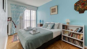 Een slaapkamer met een bed, een boekenkast, een raam met gordijnen en een aan de muur hangend schilderij.