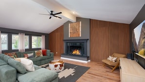 A modern living room with a fireplace, green sofa, wooden paneling, and a ceiling fan.