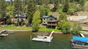 A lakeside property with a dock, a house with a deck, and a large building in the background.