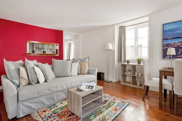 A living room with a grey sofa, a coffee table, a patterned rug, a bookshelf, and a window with curtains. - Lisbon Cozy Nest in Santa Catarina (Lisbon)