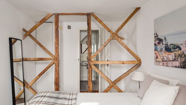 Een slaapkamer met een houten spantenplafond, een bed met witte lakens, een bedlamp en een schilderij van een stadsgezicht.